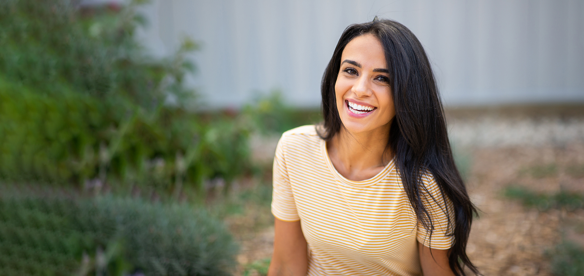 The image shows a woman with long hair smiling at the camera, wearing a yellow top and standing outdoors in front of greenery.