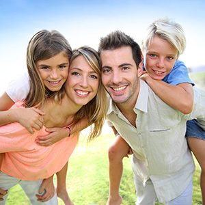 A family of four posing for a photo outdoors, with the father holding two children and the mother smiling behind them.