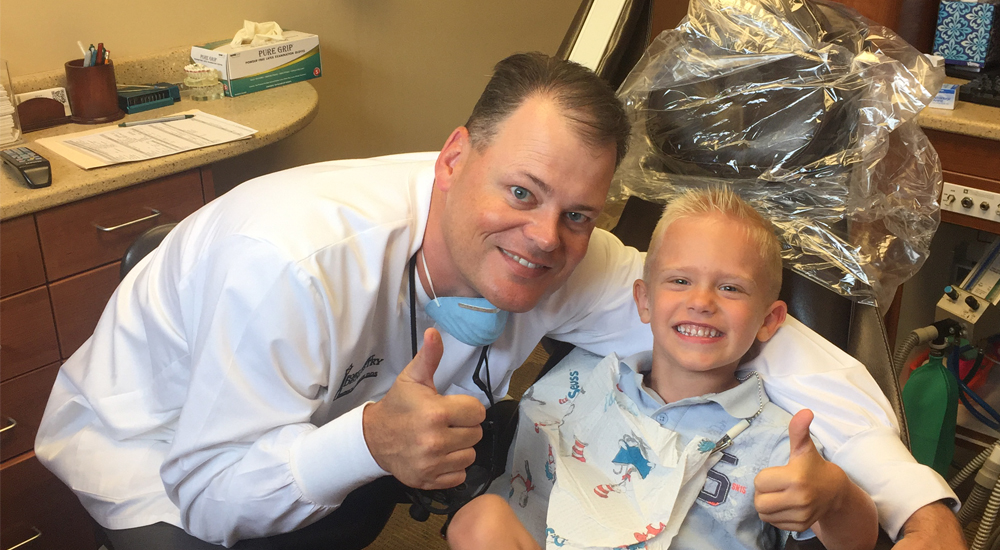The image shows a man in a white lab coat, smiling and giving a thumbs-up sign, posing with a young boy who is in a dental chair. They appear to be in an office setting, possibly a dentist s office, as suggested by the dental equipment around them.